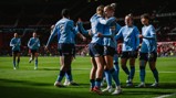 The image depicts Manchester City Women's team celebrating on the football field, wearing light blue jerseys. The stadium is filled with spectators in the background.