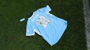 A Manchester City jersey with the name 'GUARDIOLA' and number '10' on the back, featuring images inside the number. The jersey is light blue with white details and is placed on a grassy surface.