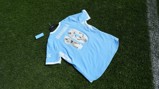 A Manchester City jersey with the name 'GUARDIOLA' and number '10' on the back, featuring images inside the number. The jersey is light blue with white details and is placed on a grassy surface.