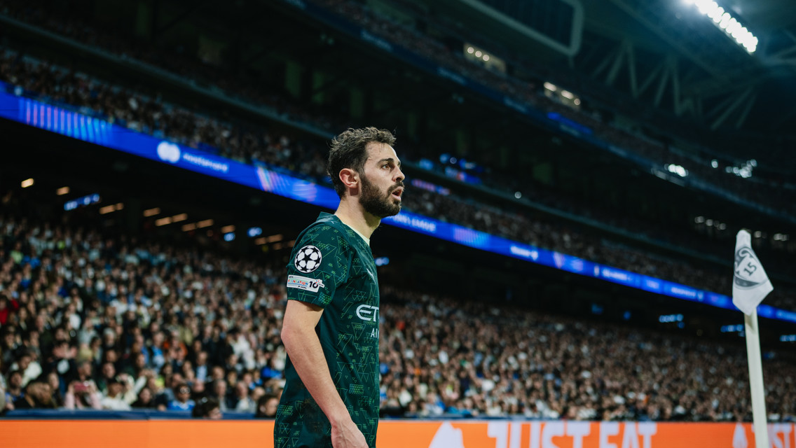 A football player in a green Manchester City jersey stands near a corner flag during a match at a stadium with a large crowd.