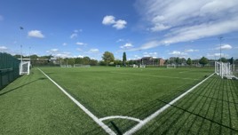 A soccer field with artificial grass, marked with white lines and goals on a clear day with blue skies and clouds.