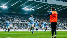 Soccer players in sky blue jerseys from Manchester City running toward the sideline celebrating a goal in a packed stadium, with 'UNITED' visible on the roof structure.