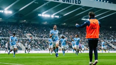 Soccer players in sky blue jerseys from Manchester City running toward the sideline celebrating a goal in a packed stadium, with 'UNITED' visible on the roof structure.