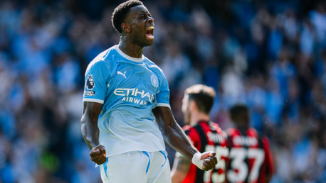 A Manchester City player passionately celebrating a goal in a match. He is wearing a blue Manchester City jersey with clenched fists and a shout of triumph, while other players are seen in the background.