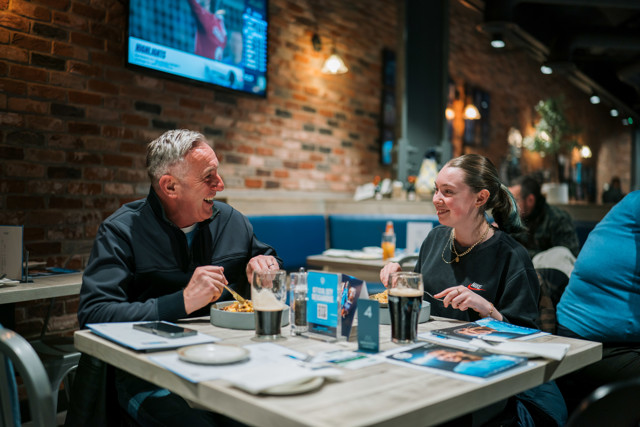 Two people enjoying a meal and conversation at a restaurant table with food and drinks. There is a television on the wall showing sports.
