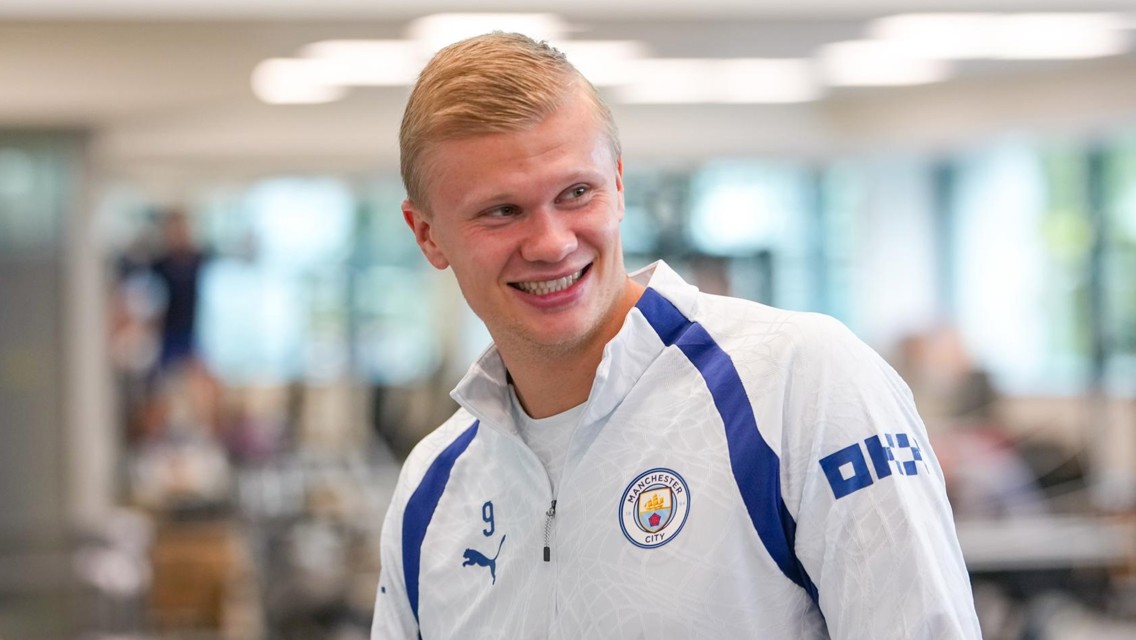 A person wearing a Manchester City tracksuit top smiling indoors.