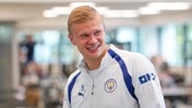 A person wearing a Manchester City tracksuit top smiling indoors.