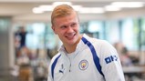 A person wearing a Manchester City tracksuit top smiling indoors.