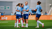 Players wearing light blue jerseys celebrating on a soccer field. One player has number 9, another has number 8, and another has number 57 on the back of their jerseys. They are smiling and appear to be from Manchester City Women’s Team.