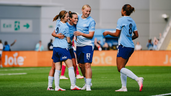 Players wearing light blue jerseys celebrating on a soccer field. One player has number 9, another has number 8, and another has number 57 on the back of their jerseys. They are smiling and appear to be from Manchester City Women’s Team.