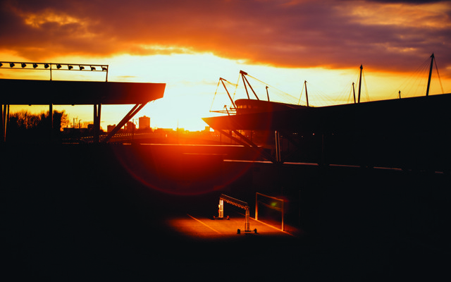 Silhouette of a football stadium with the sun setting in the background, casting an orange glow over the scene.
