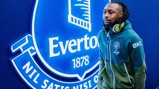 A person wearing Manchester City apparel stands beside an illuminated Everton logo display.