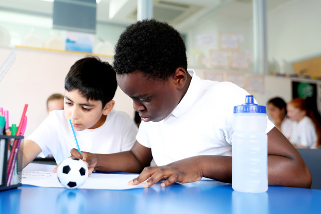 Two students sitting at a classroom desk working on an assignment. One student holds a pencil while the other focuses on the paper. A small soccer ball and a water bottle are on the desk.