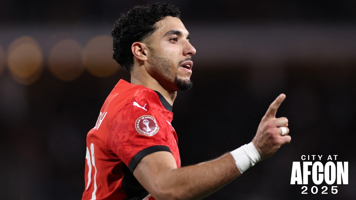 A football player wearing a red Puma jersey raises one finger, signifying celebration, with AFCON 2025 logo visible on sleeve.