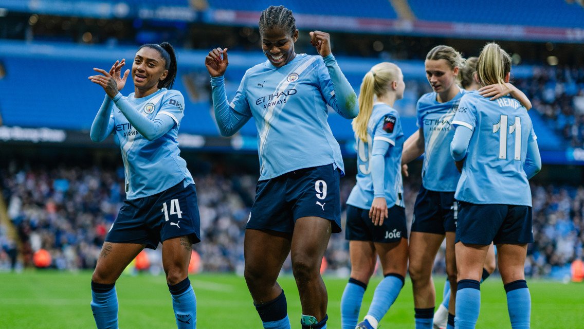 Manchester City Women's team players celebrating on the field in blue jerseys with the Etihad logo. The stadium background indicates a sports venue, possibly the Etihad Stadium.