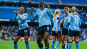 Manchester City Women's team players celebrating on the field in blue jerseys with the Etihad logo. The stadium background indicates a sports venue, possibly the Etihad Stadium.