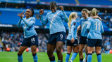Manchester City Women's team players celebrating on the field in blue jerseys with the Etihad logo. The stadium background indicates a sports venue, possibly the Etihad Stadium.