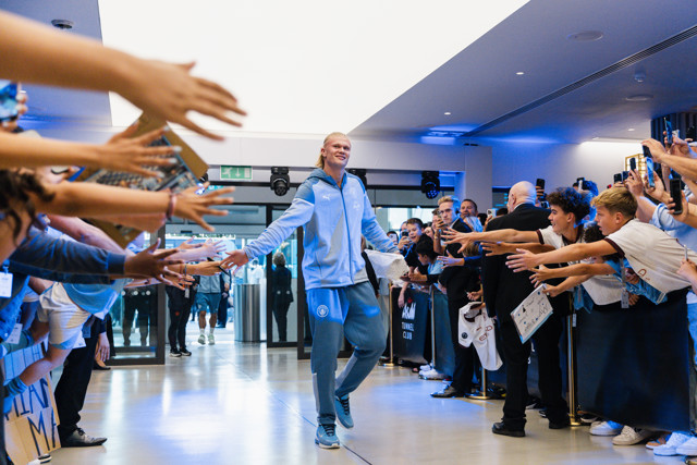 A male individual wearing a Manchester City blue tracksuit walks through a crowd of fans, high-fiving them. The scene takes place in the Asahi Tunnel Club, with fans wearing light blue jerseys and holding a football with a red cross.