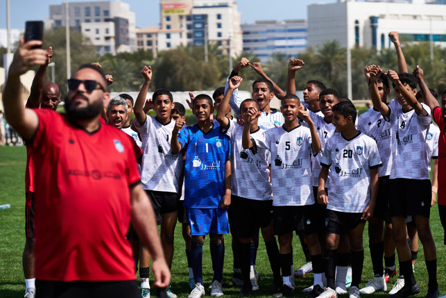 Focused on a group of young soccer players in white shirts standing on a field raising their hands. One person in the front is taking a selfie.