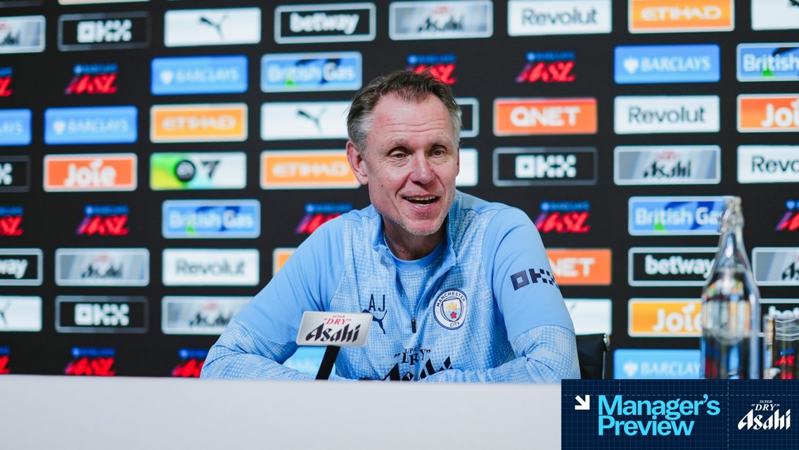 A person is speaking at a press conference wearing a Manchester City jacket with sponsors visible on the backdrop.