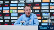 A person is speaking at a press conference wearing a Manchester City jacket with sponsors visible on the backdrop.