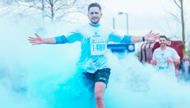 A runner wearing a white T-shirt with number 1489 runs through a cloud of blue powder, spreading arms wide. Another runner is visible in the background, partially obscured by the blue powder.