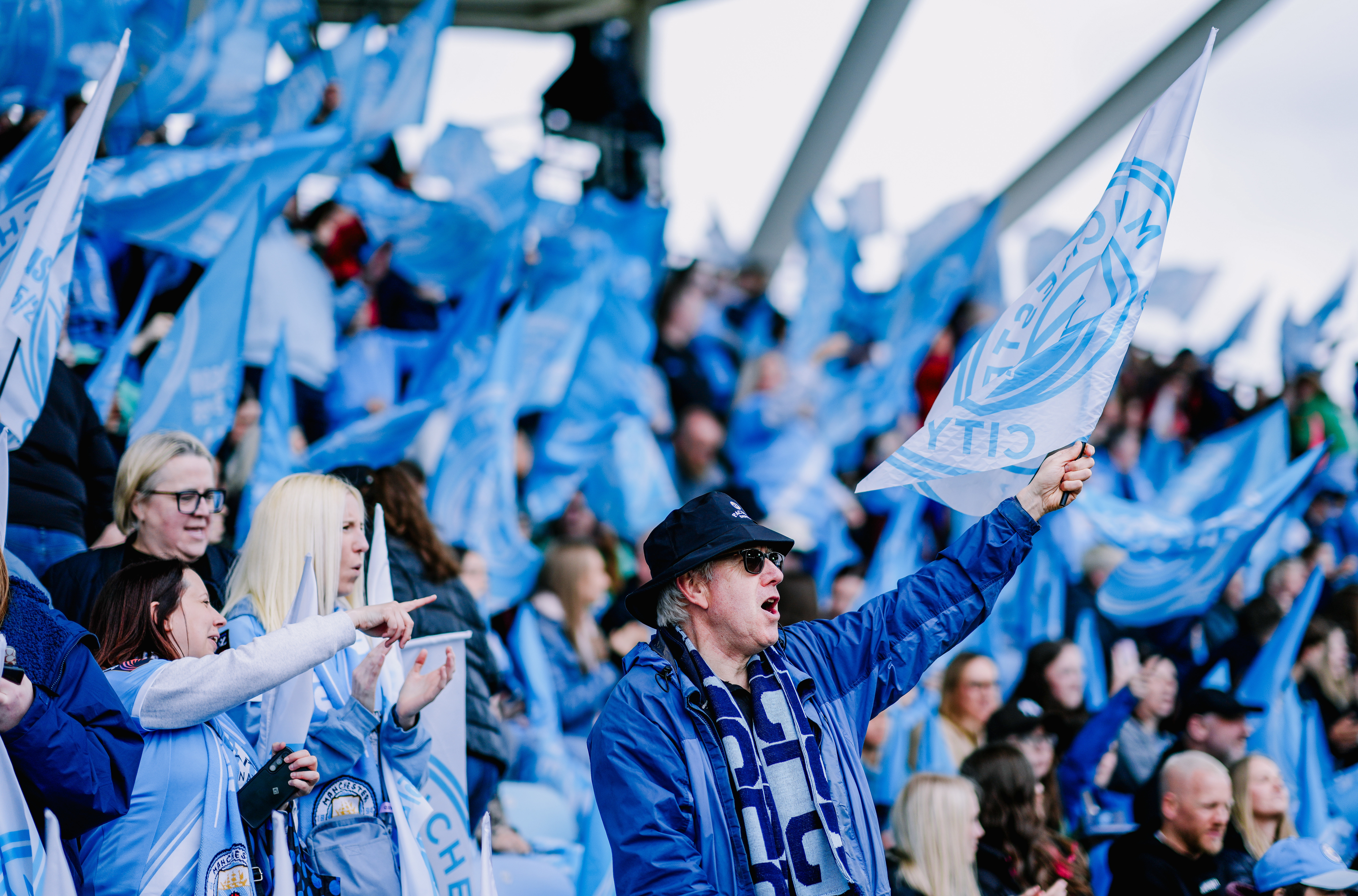 A crowd of Manchester City fans waving blue flags during a sports event at a stadium.