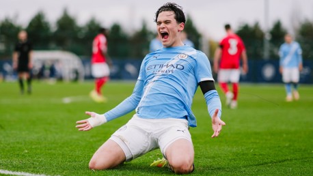 A football player wearing a light blue Manchester City kit is kneeling on the field in celebration, with players from the opposing team in red in the background.
