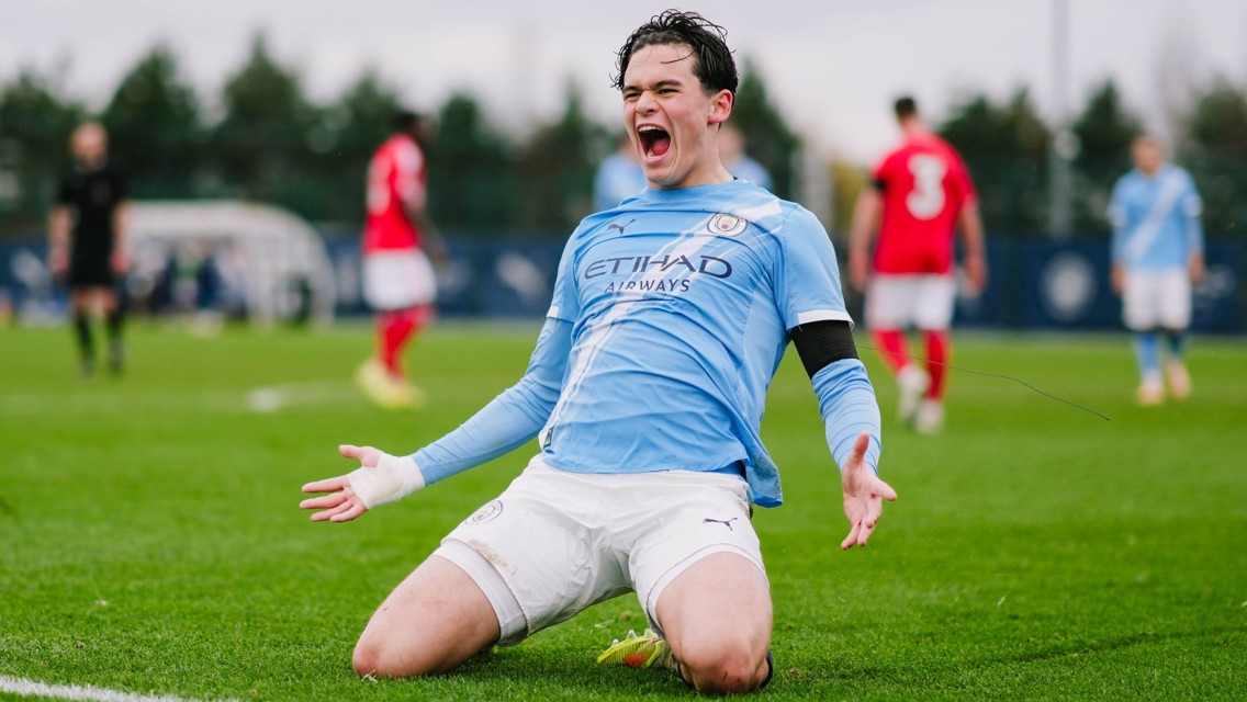 A football player wearing a light blue Manchester City kit is kneeling on the field in celebration, with players from the opposing team in red in the background.
