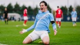 A football player wearing a light blue Manchester City kit is kneeling on the field in celebration, with players from the opposing team in red in the background.