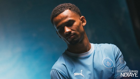 A football player in a light blue Manchester City jersey stands against a gradient background. The text 'Welcome to City' and 'Iliman Ndiaye' appears alongside a Puma logo on the shirt.