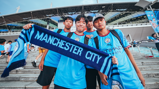A group of four fans in Manchester City jerseys hold a scarf outside a stadium entrance. They appear enthusiastic and are part of a larger crowd attending an event.