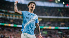 A Manchester City player celebrating a goal in a football match. The player is wearing a light blue Manchester City jersey.