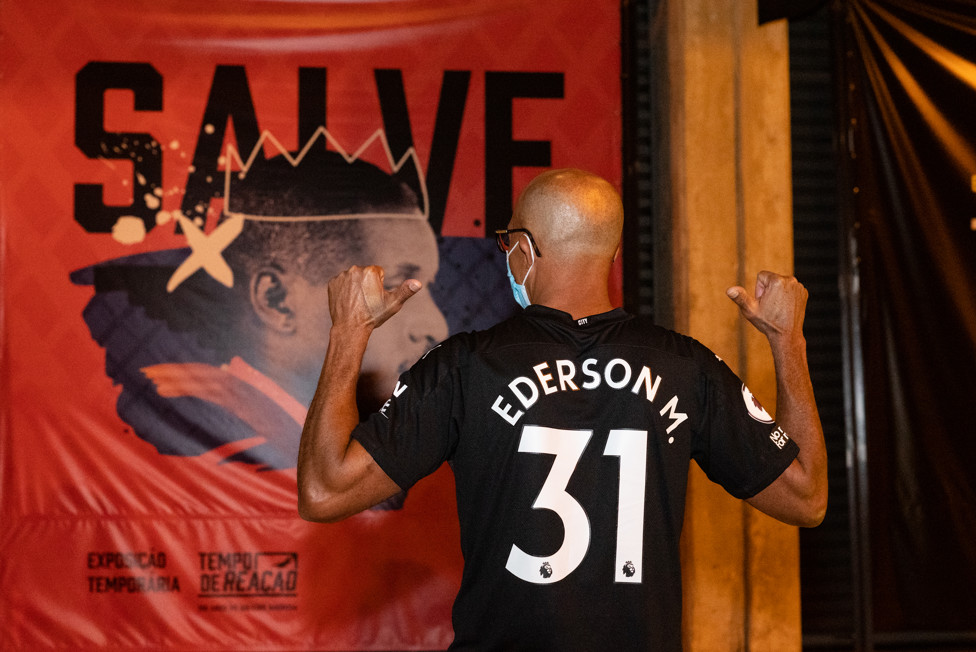 Bartolomeu Moraes, pai de Ederson, posa ao lado de banner do goleiro Barbosa no Museu do Futebol