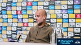 A blurred-faced individual, possibly a football manager, sitting at a press conference with a backdrop displaying Manchester City sponsors' logos. Microphone and bottled water visible on the table.
