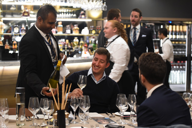 A waiter pours wine for a guest at a formal dinner event with multiple tables set with glassware and cutlery. Background shows a bar area with more people and various bottles on display.