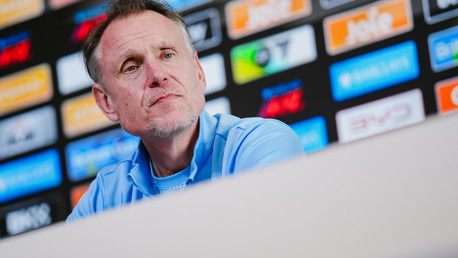 A person in a light blue outfit is seated at a press conference table with various logos in the background.