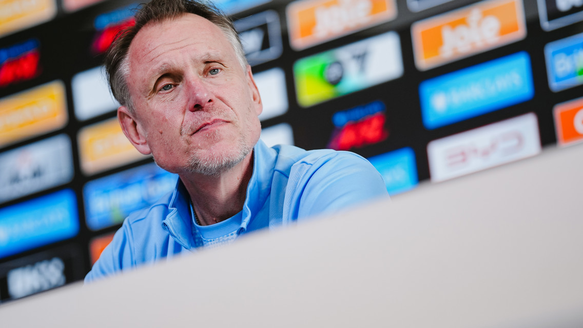 A person in a light blue outfit is seated at a press conference table with various logos in the background.