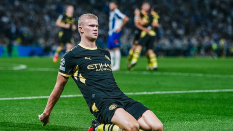 A Manchester City player in a black and gold kit slides on his knees, celebrating a goal during a match.