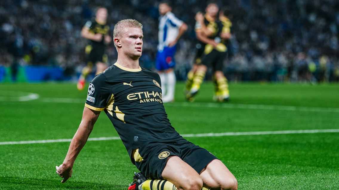 A Manchester City player in a black and gold kit slides on his knees, celebrating a goal during a match.
