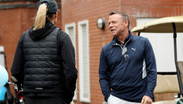 Two individuals in golf attire, one man in a blue Puma jacket and one woman in a black vest, conversing outdoors next to a golf cart. The man's face is blurred.
