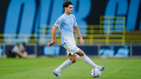 A soccer player wearing a light blue Manchester City jersey is in action on the field, controlling the ball with his left foot.