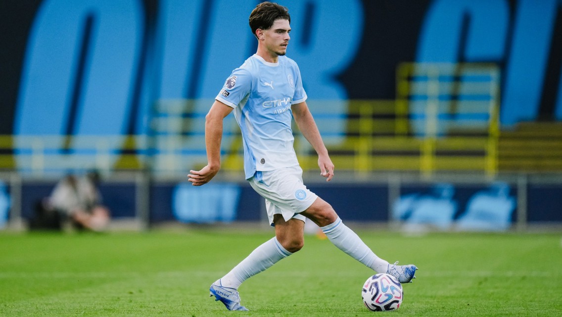 A soccer player wearing a light blue Manchester City jersey is in action on the field, controlling the ball with his left foot.