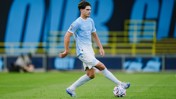 A soccer player wearing a light blue Manchester City jersey is in action on the field, controlling the ball with his left foot.