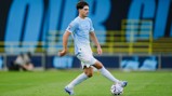 A soccer player wearing a light blue Manchester City jersey is in action on the field, controlling the ball with his left foot.