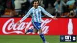 A football player in an Argentina jersey celebrating with arms outstretched on the field. The background features stadium advertising and city at the world cup branding.