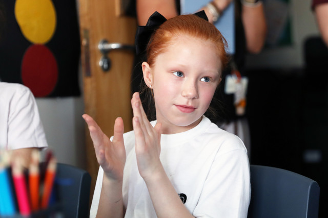A child with red hair tied with a black ribbon claps their hands, with colorful pencils in the foreground and a blurred background including a door and partially visible people.