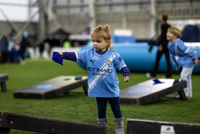 A young child wearing a Manchester City jersey participates in an indoor event, playing with bean bags on a grassy pitch.
