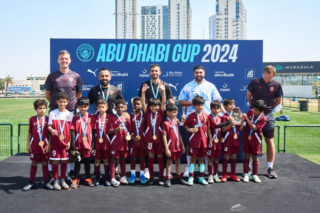 A youth soccer team with coaches and officials posing for a group photo after winning the Abu Dhabi Cup 2024. The players, wearing maroon jerseys, hold medals and a trophy. A banner in the background reads 'Abu Dhabi Cup 2024' with sponsor logos.