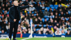 A bald coach in a dark jacket gestures and points from the sideline during a game with a blurred crowd in the background.